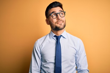 Young handsome businessman wearing tie and glasses standing over yellow background smiling looking...