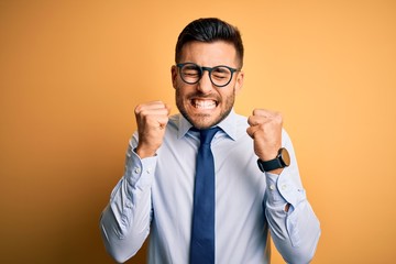 Young handsome businessman wearing tie and glasses standing over yellow background excited for success with arms raised and eyes closed celebrating victory smiling. Winner concept.