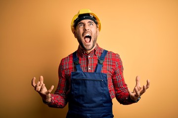 Young builder man wearing construction uniform and safety helmet over yellow isolated background crazy and mad shouting and yelling with aggressive expression and arms raised. Frustration concept.