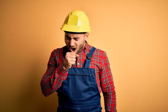 Young Builder Man Wearing Construction Uniform And Safety Helmet Over Yellow Isolated Background Feeling Unwell And Coughing As Symptom For Cold Or Bronchitis. Health Care Concept.