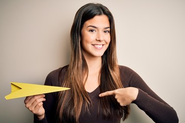 Young beautiful girl holding paper plane standing over isolated white background with surprise face pointing finger to himself