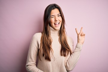 Young beautiful girl wearing casual turtleneck sweater standing over isolated pink background smiling with happy face winking at the camera doing victory sign. Number two.