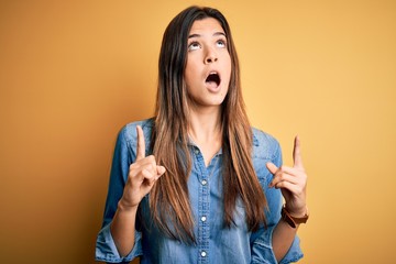 Fototapeta premium Young beautiful girl wearing casual denim shirt standing over isolated yellow background amazed and surprised looking up and pointing with fingers and raised arms.