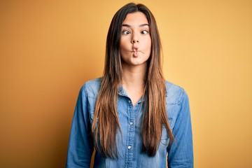 Fototapeta premium Young beautiful girl wearing casual denim shirt standing over isolated yellow background making fish face with lips, crazy and comical gesture. Funny expression.