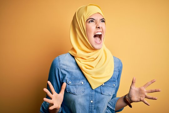 Young beautiful girl wearing muslim hijab standing over isolated yellow background crazy and mad shouting and yelling with aggressive expression and arms raised. Frustration concept.