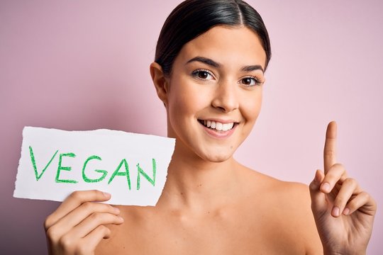 Young beautiful girl holding paper with vegan message over isolated pink background surprised with an idea or question pointing finger with happy face, number one