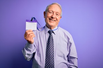 Senior grey haired business man holding identification tag over purple background with a happy face standing and smiling with a confident smile showing teeth