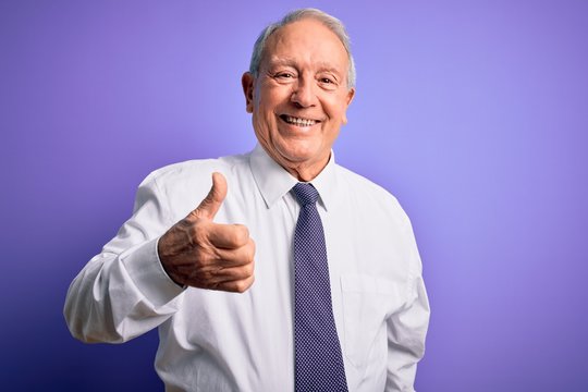 Grey haired senior business elegant man standing over purple isolated background doing happy thumbs up gesture with hand. Approving expression looking at the camera showing success.