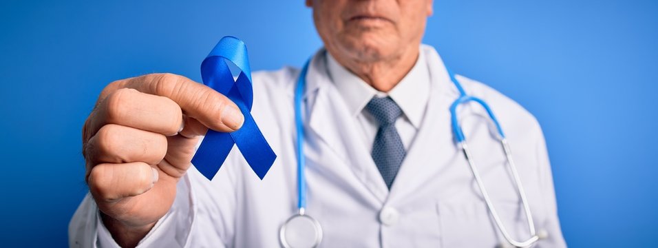 Grey haired senior doctor man holding colon cancer awareness blue ribbon over blue background with a confident expression on smart face thinking serious