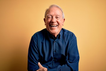 Grey haired senior man wearing casual blue shirt standing over yellow background smiling and laughing hard out loud because funny crazy joke with hands on body.