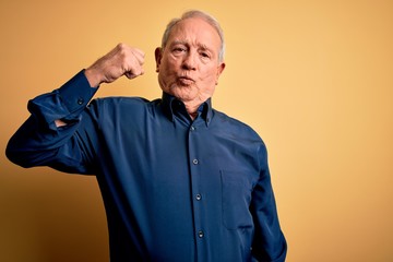 Grey haired senior man wearing casual blue shirt standing over yellow background Strong person...