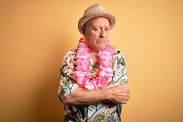 Grey haired senior man wearing summer hat and hawaiian lei over yellow background skeptic and...