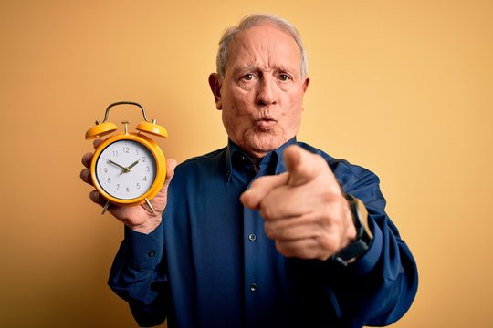 Senior grey haired man holding vintage alarm clock over yellow background pointing with finger to the camera and to you, hand sign, positive and confident gesture from the front