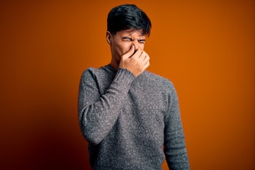 Young handsome man wearing casual sweater standing over isolated orange background smelling something stinky and disgusting, intolerable smell, holding breath with fingers on nose. Bad smell