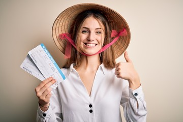 Young beautiful redhead tourist woman wearing asian traditional hat holding boarding pass happy with big smile doing ok sign, thumb up with fingers, excellent sign