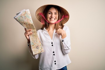 Young beautiful redhead woman wearing asian traditional hat holding city map with surprise face pointing finger to himself