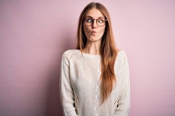 Obraz premium Young beautiful redhead woman wearing casual sweater and glasses over pink background making fish face with lips, crazy and comical gesture. Funny expression.