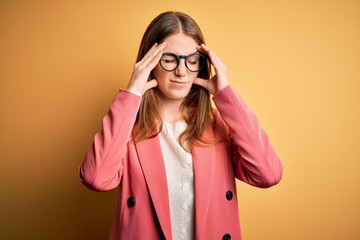 Young beautiful redhead woman wearing jacket and glasses over isolated yellow background with hand on head for pain in head because stress. Suffering migraine.