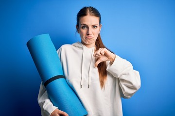 Young beautiful redhead sporty woman doing sport holding yoga mat over blue background with angry...