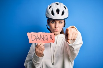 Young beautiful redhead cyclist woman wearing bike helmet holding danger paper message pointing with finger to the camera and to you, hand sign, positive and confident gesture from the front