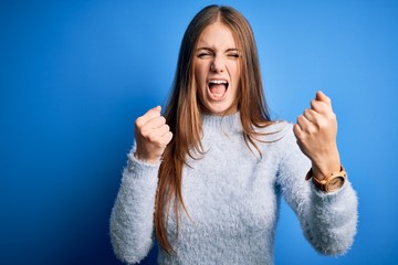 Young beautiful redhead woman wearing casual sweater over isolated blue background angry and mad raising fists frustrated and furious while shouting with anger. Rage and aggressive concept.