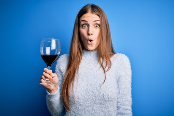 Young beautiful redhead woman drinking glass of red wine over isolated blue background scared in...