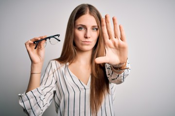 Obraz premium Young beautiful redhead optical woman holding glasses over isolated white background with open hand doing stop sign with serious and confident expression, defense gesture