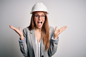 Young beautiful redhead architect woman wearing security helmet over white background celebrating crazy and amazed for success with arms raised and open eyes screaming excited. Winner concept
