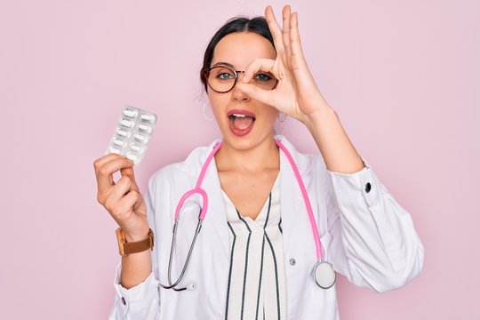 Young beautiful doctor woman with blue eyes wearing stethoscope holding medicine pills with happy face smiling doing ok sign with hand on eye looking through fingers