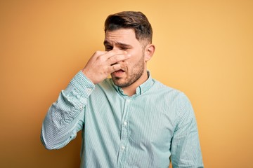 Young business man with blue eyes wearing elegant green shirt over yellow background smelling something stinky and disgusting, intolerable smell, holding breath with fingers on nose. Bad smell