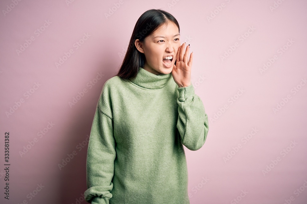 Poster Young beautiful asian woman wearing green winter sweater over pink solated background shouting and screaming loud to side with hand on mouth. Communication concept.