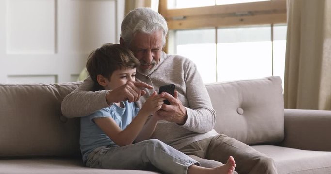 Smart little child boy teaching elderly mature grandfather using applications on smartphone. Happy middle aged man cuddling small grandson, spending free leisure time at home, web surfing information.