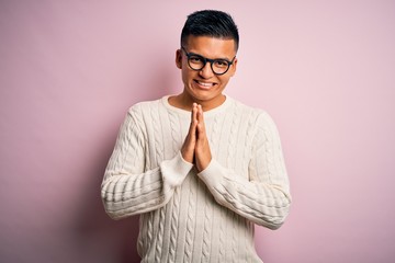 Young handsome latin man wearing white casual sweater and glasses over pink background praying with hands together asking for forgiveness smiling confident.