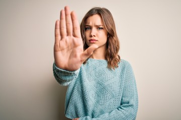 Young blonde girl wearing casual blue winter sweater over isolated background doing stop sing with palm of the hand. Warning expression with negative and serious gesture on the face.