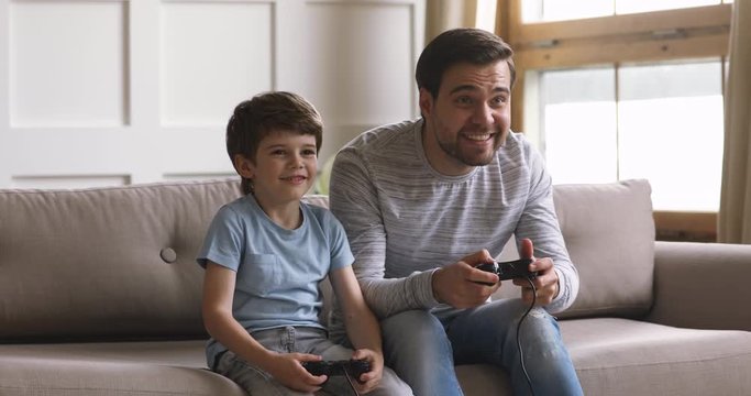 Overjoyed little school aged boy resting on sofa with handsome emotional father, playing online video game at home. Excited two generations family enjoying weekend playtime together.