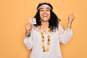African american curly hippie woman wearing sunglasses holding reminder with peace symbol screaming...