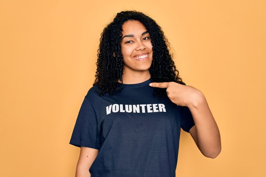Young african american curly woman doing volunteering wearing volunteer t-shirt with surprise face pointing finger to himself
