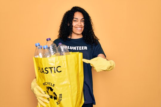 Young african american woman doing volunteering recycling holding bag with plastic bottles very happy pointing with hand and finger