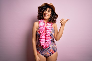 Young beautiful arab woman on vacation wearing swimsuit and hawaiian lei flowers smiling with happy face looking and pointing to the side with thumb up.