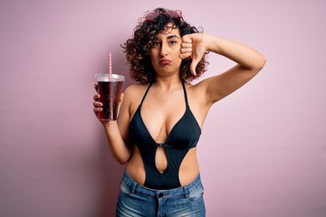 Beautiful arab woman on vacation wearing swimsuit drinking cola refreshment using straw with angry face, negative sign showing dislike with thumbs down, rejection concept