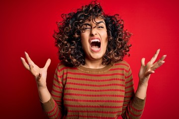 Young beautiful curly arab woman wearing casual striped sweater standing over red background crazy and mad shouting and yelling with aggressive expression and arms raised. Frustration concept.