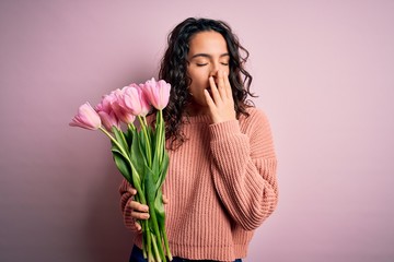 Young beautiful romantic woman with curly hair holding bouquet of pink tulips bored yawning tired...