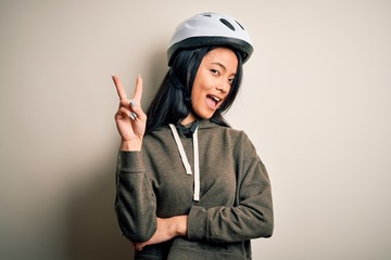 Young beautiful chinese woman wearing bike helmet over isolated white background smiling with happy face winking at the camera doing victory sign. Number two.