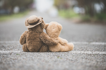 One teddy bear with his/her arm wrapped around a smaller teddy bear showing compassion on a road - obrazy, fototapety, plakaty