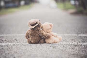 One teddy bear with his/her arm wrapped around a smaller teddy bear showing compassion on a paved...