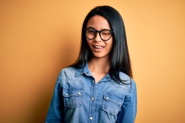 Young beautiful chinese woman wearing casual denim shirt over isolated yellow background winking...