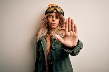 Young african american skier woman with curly hair wearing snow sportswear and ski goggles doing stop sing with palm of the hand. Warning expression with negative and serious gesture on the face.