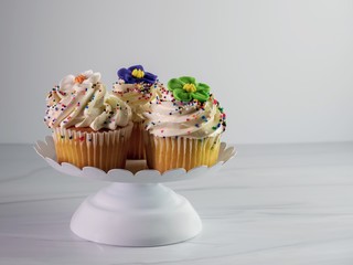 3 cupcakes with white frosting, fondant flower and sprinkles on a white platter with a sandstone background.  Festive, delicious and perfect for a celebration.