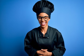 Young african american chef woman wearing cooker uniform and hat over blue background smiling and laughing hard out loud because funny crazy joke with hands on body.