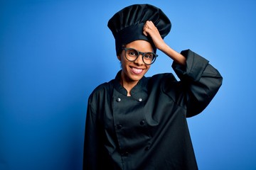 Young african american chef woman wearing cooker uniform and hat over blue background smiling confident touching hair with hand up gesture, posing attractive and fashionable